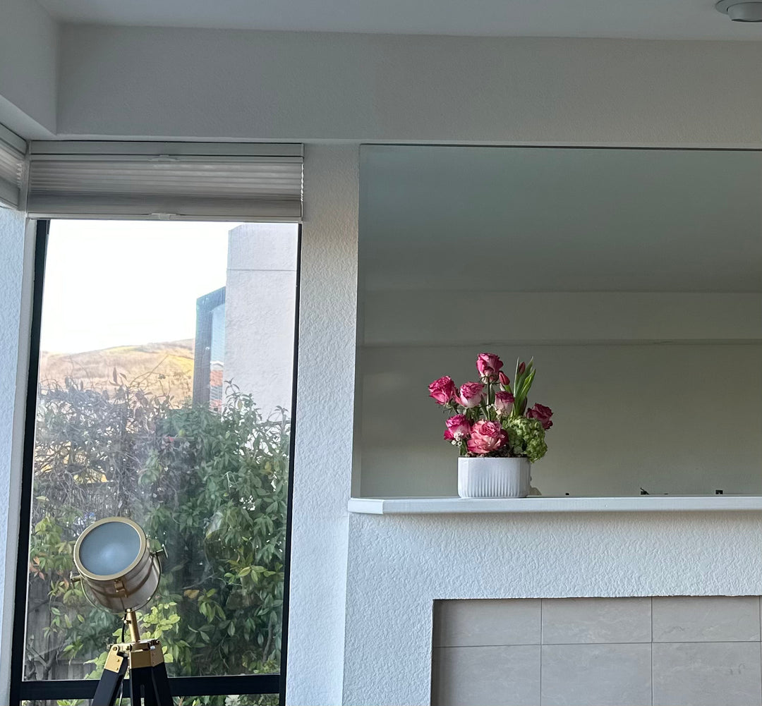 Floral arrangement on a shelf with a view of trees outside a window.