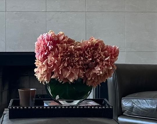 A large bouquet of ruffled pink flowers arranged in a clear glass vase on a black tray, placed in a modern interior setting with a tiled wall and dark leather chair visible.