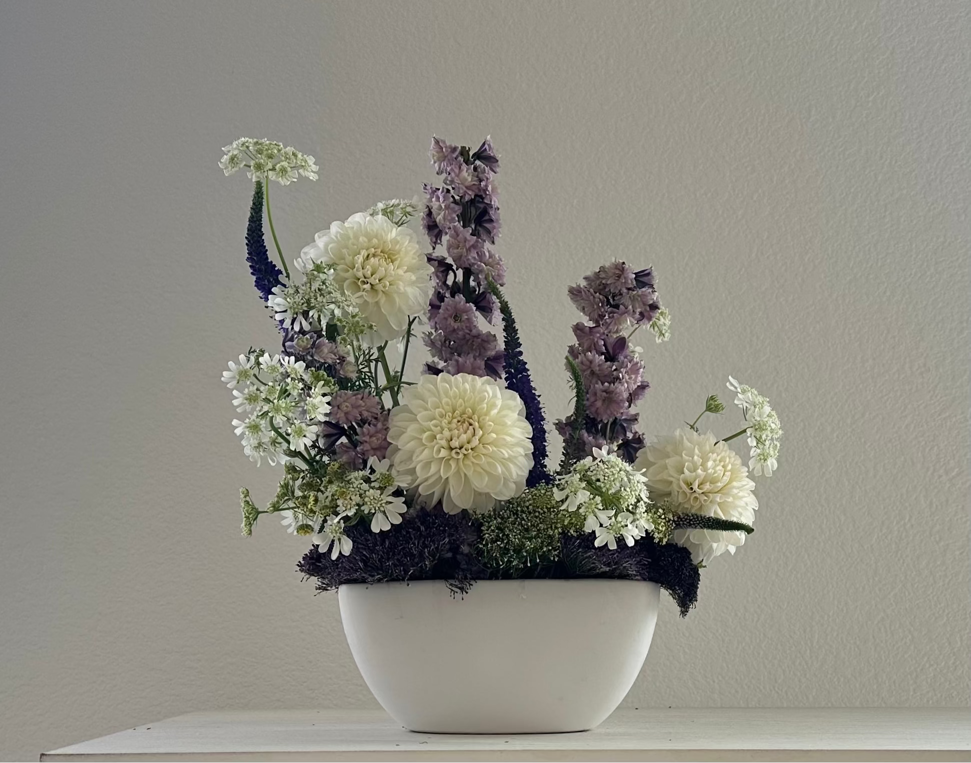 Floral arrangement in a white ceramic bowl, featuring white and lavender-purple flowers with green foliage, set against a plain light gray wall.
